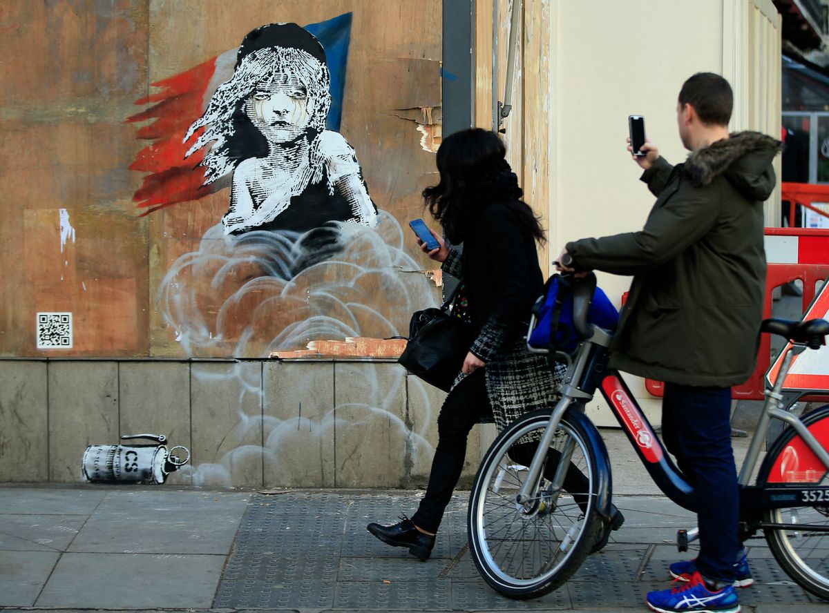 Commuters take photos on their phones of a new artwork by British artist Banksy opposite the French Embassy, in London, Monday, Jan. 25, 2016. The artwork depicts the young girl from the musical Les Miserables with tears streaming from her eyes as a can of CS gas lies beneath her. The work is criticising the use of teargas in the refugee camp in Calais. (AP Photo/Alastair Grant) (AP)