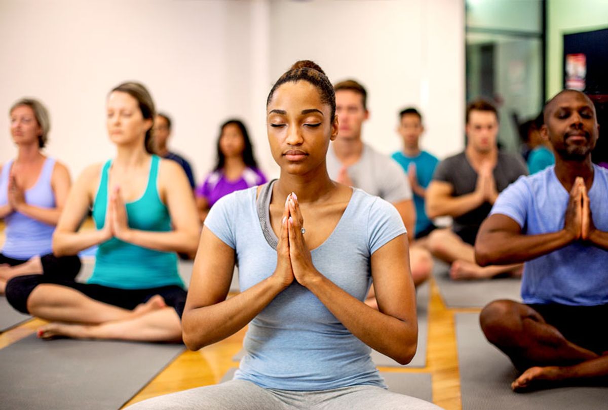 A multi-ethnic group of adults are taking a yoga class together at the gym. They are sitting on their exercise mats and are meditating with their eyes closed and their hands together at heart center. (Getty Images)