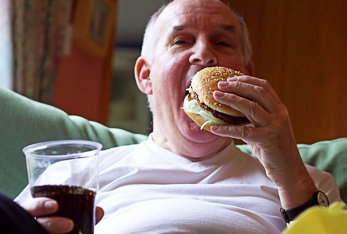 Senior man on sofa, eating hamburger (Getty Images)