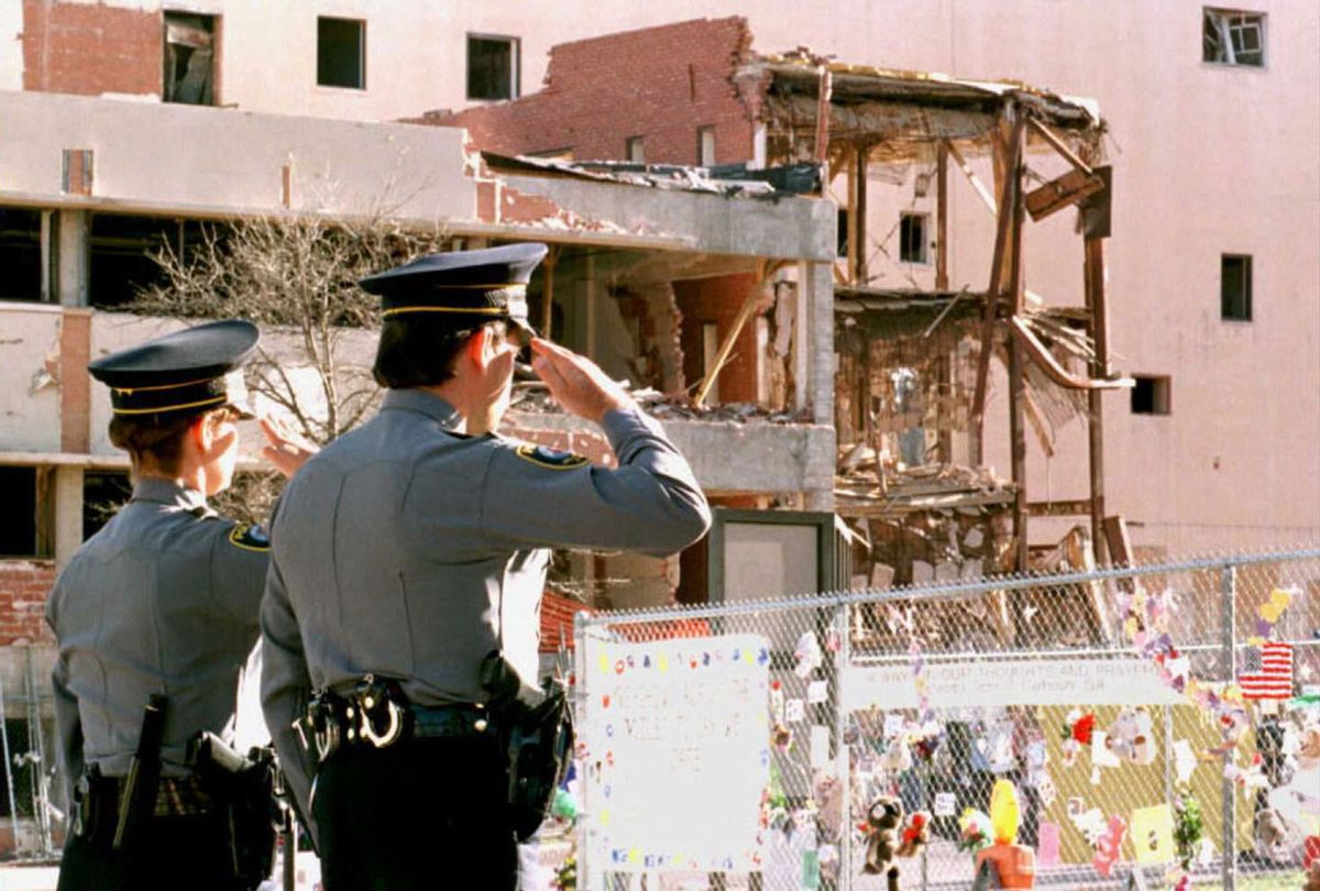 Oklahoma City police officers salute 19 April during a moment of silence at a memorial service held on the first anniversary of the bombing of the Alfred P. Murrah Federal Building as they face the fence surrounding the bomb site. In the background are buildings destroyed in the blast that killed 168 adults and children. (CHRIS WILKINS/AFP via Getty Images)