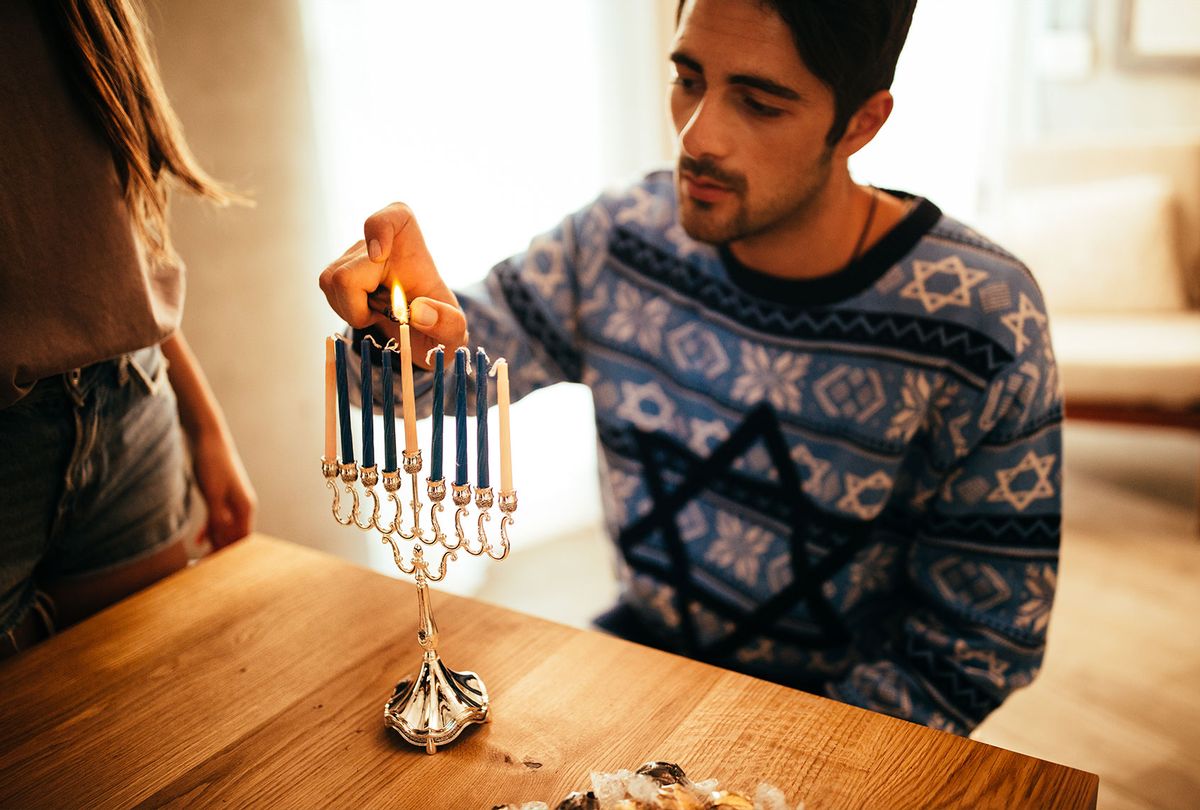 Man wearing a Hanukkah sweater lights a Hanukkah menorah  (Getty Image/wundervisuals)