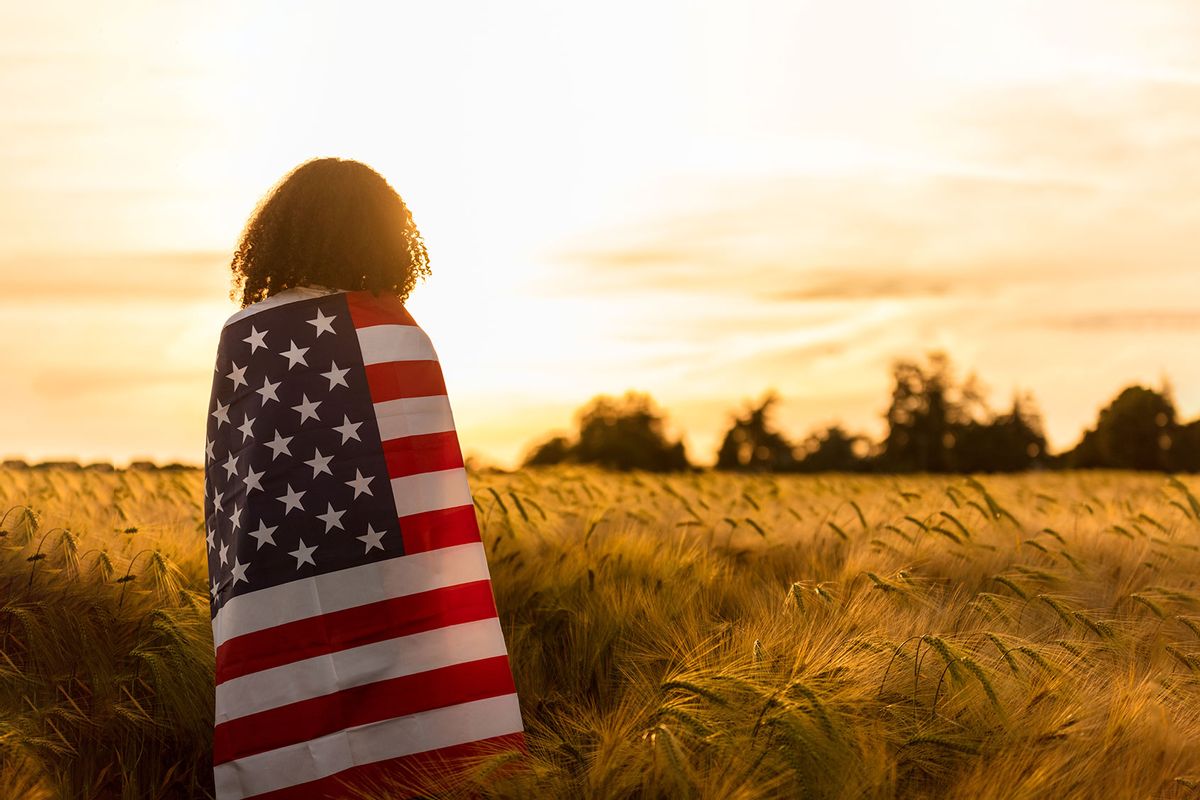 African American girl teenager female young woman in a field of wheat or barley crops wrapped in USA stars and stripes flag in golden sunset evening sunshine (Getty Images/dmbaker)