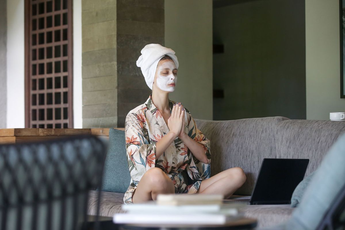 Young woman sitting on sofa with face beauty mask in front of laptop and doing yoga or meditation (Getty Images/triocean)