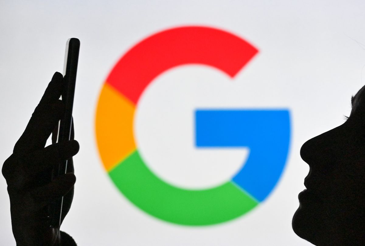 A woman holding a cell phone in front of the Google logo displayed on a computer screen.  (Artur Widak/NurPhoto via Getty Images)