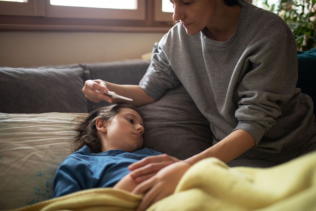 Boy lying sick on the sofa at home while his mother takes care of him and helping him to measure his temperature with a digital thermometer. (Getty Images/miljko)