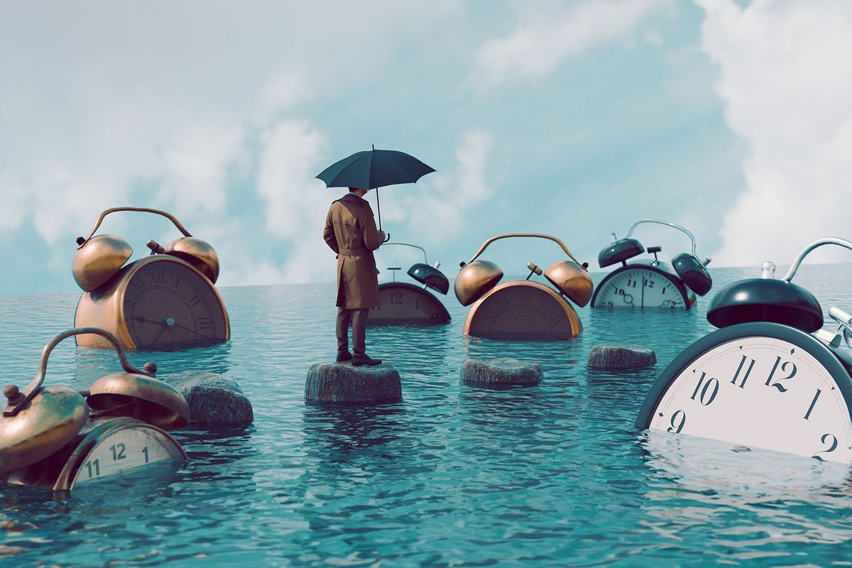 Man stands with umbrella outside looking at large collection of big alarm clocks drowning in the sea. (Getty Images/mikkelwilliam)