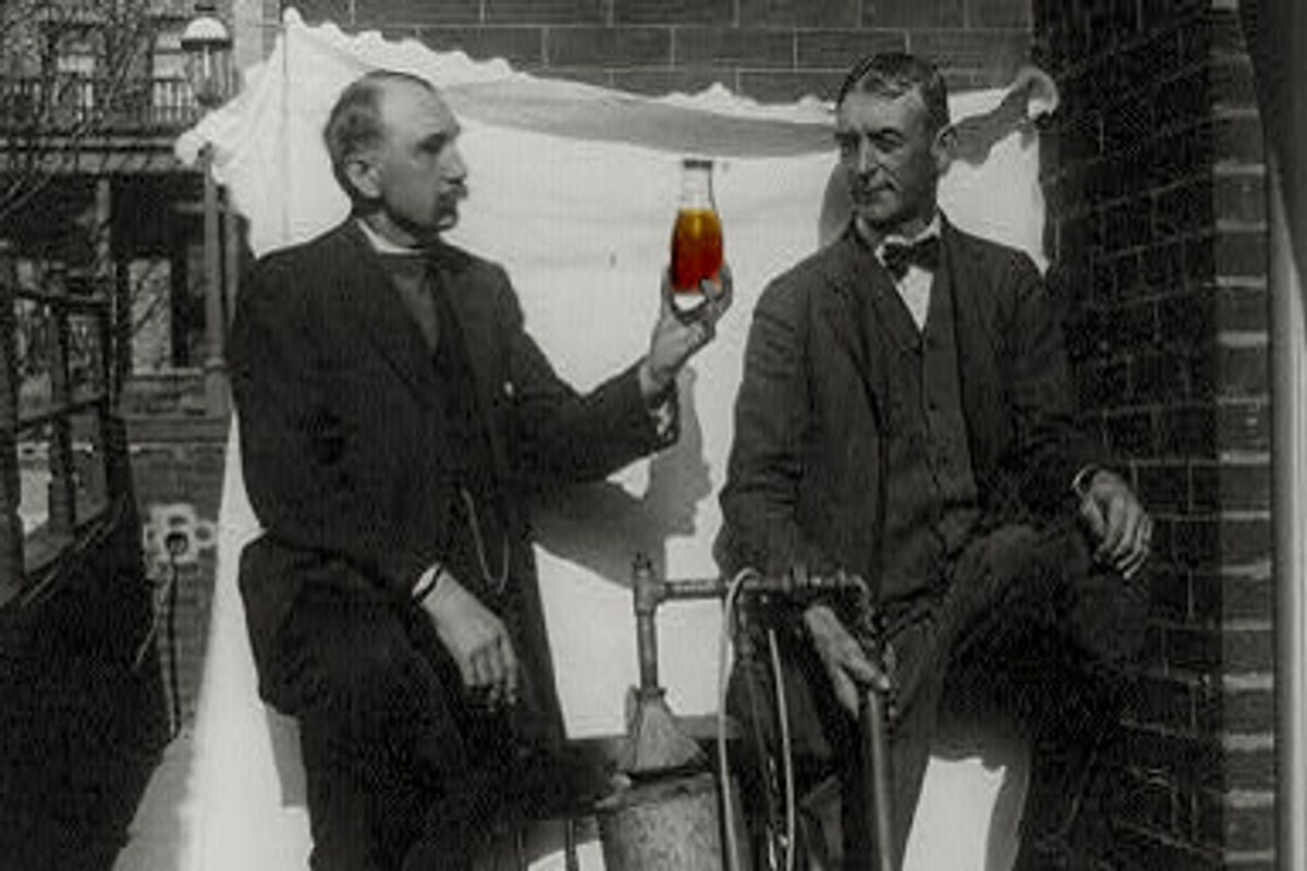Two men standing outdoors with small still, one of them holding up bottle of liquor.