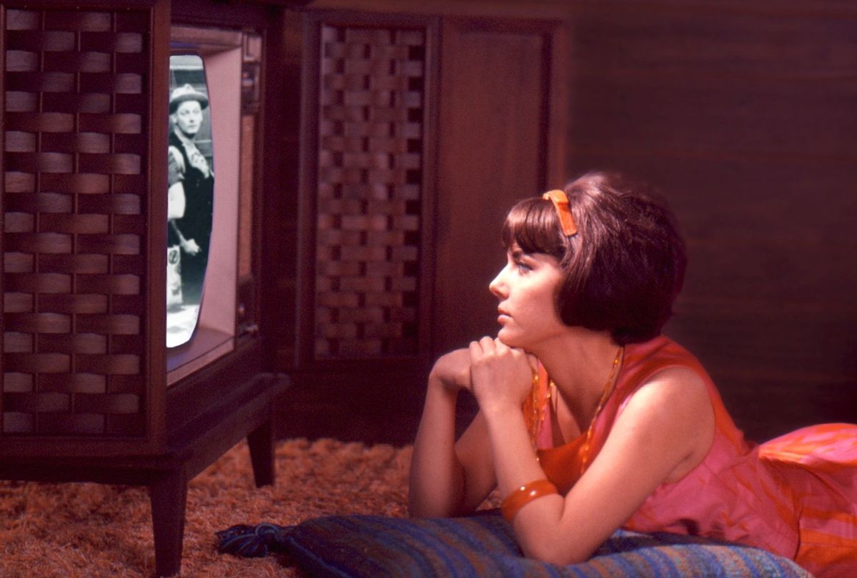 A composite image shows a young woman in a pink and orange dress lies on the floor and watches an episode of 'The Honeymooners' on a console television set, late 1960s or early 1970s. On the screen is an image of American actor Art Carney (1918 - 2003) (Tom Kelley/Getty Images)
