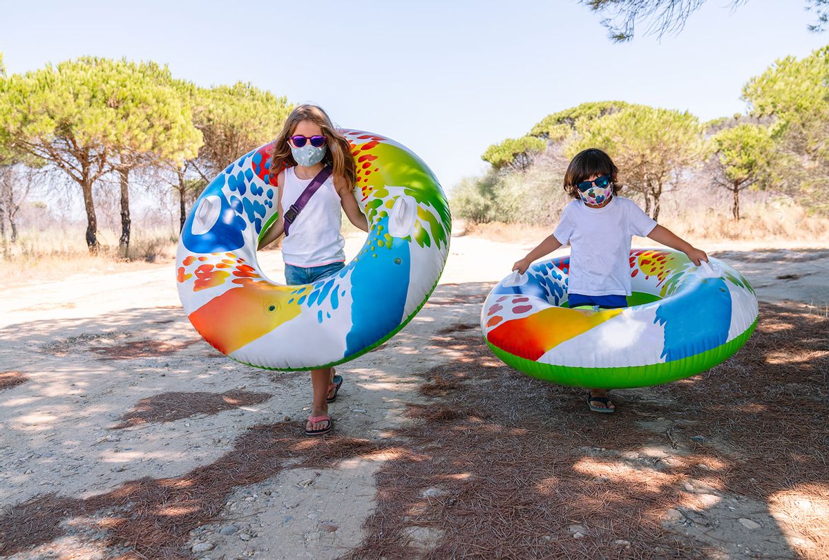 Two children with a mask on their face and glasses walking through a campground with a colorful inflatable pool tubes (Getty Images)