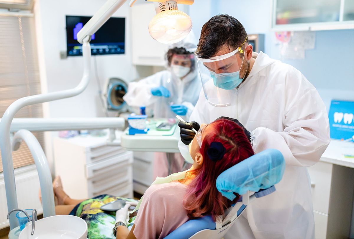 Dentist working with patient while dental assistant is helping around, both wearing a protective gowns, face masks and gloves during COVID-19 pandemic (Getty Images/Kosamtu)