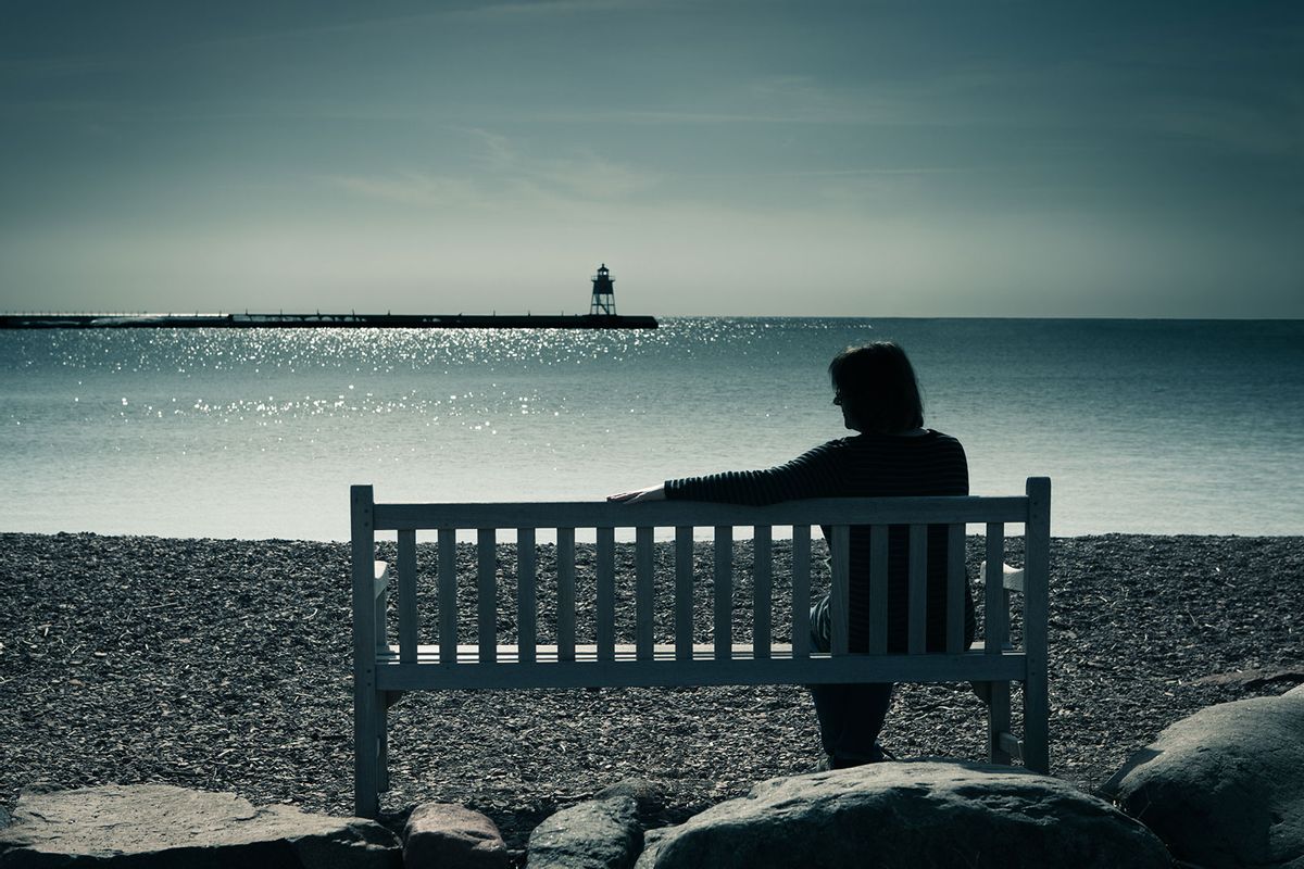 Woman in silhouette, sitting on a bench in a moody landscape at water's edge (Getty Images/YinYang)