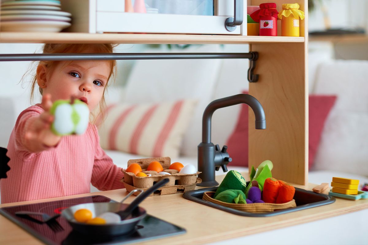 Cute toddler playing with a toy kitchen at home, roasting eggs and treating you with an apple slice. (Getty Images/olesiabilkei)