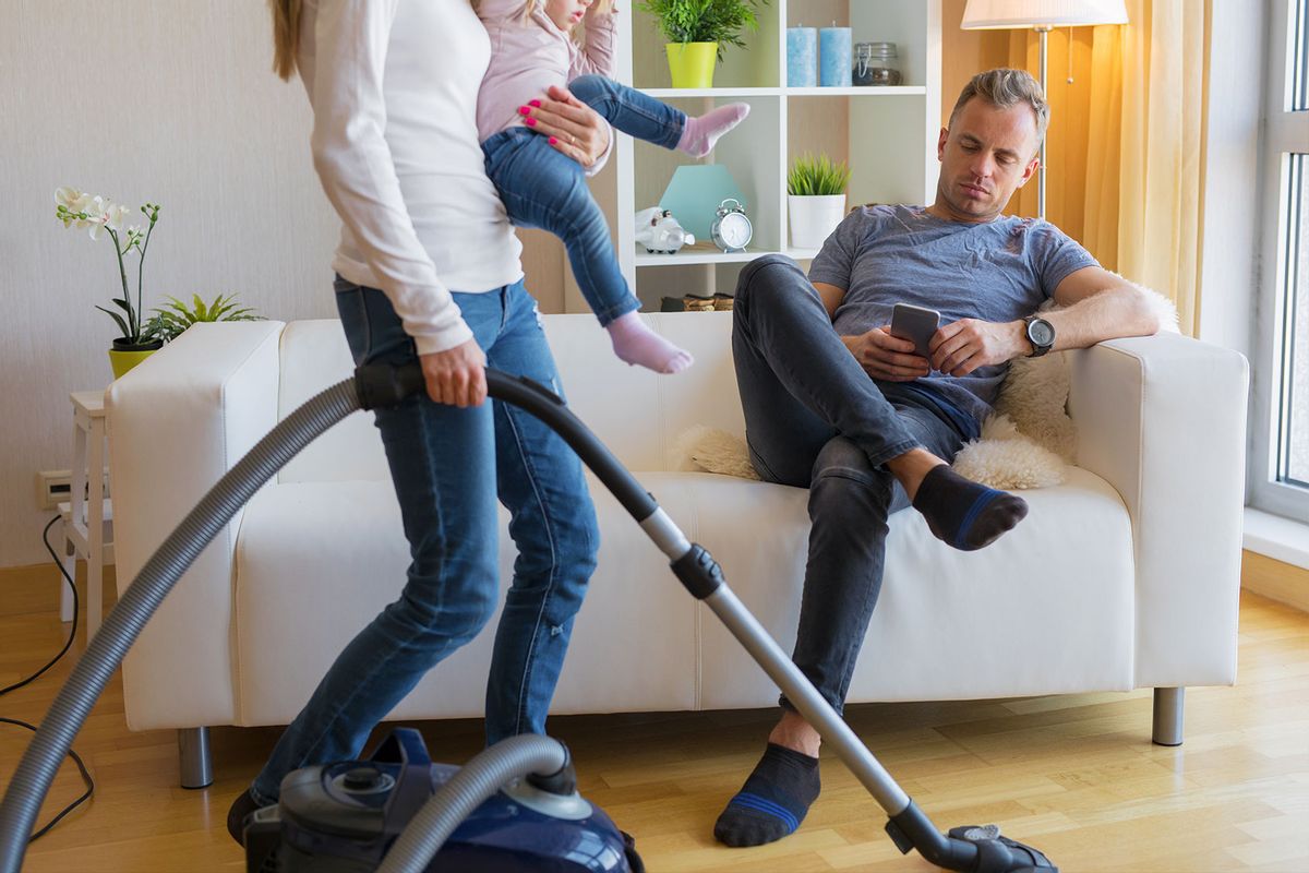 Woman with small child in her hands doing housekeeping while man sitting in couch and relaxing (Getty Images/grinvalds)