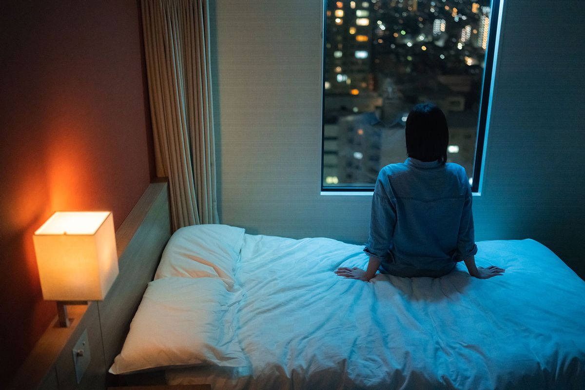 A rear view of a woman sitting alone on a bed in room and looking through the window at night. (Getty Images/recep-bg)