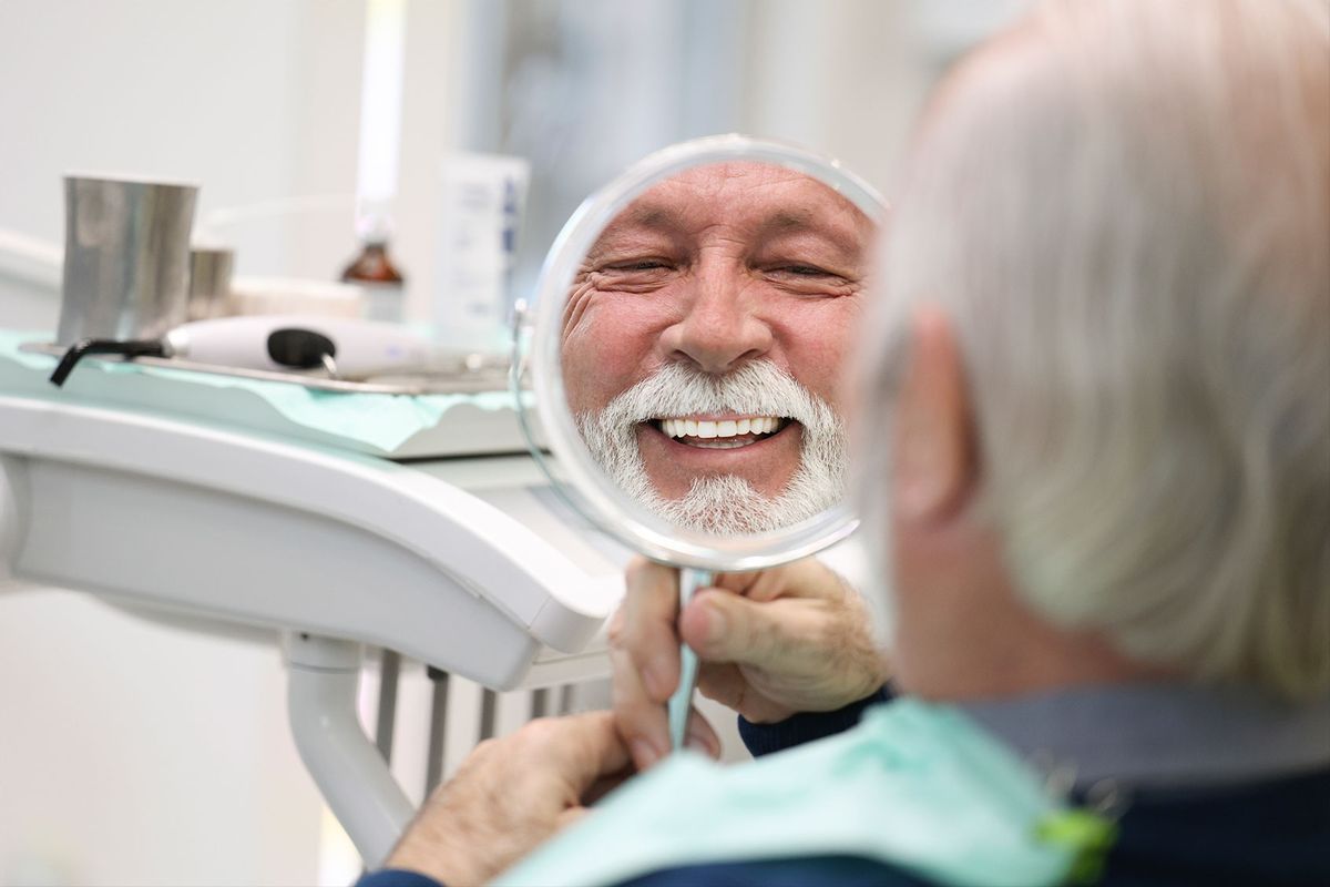 Senior man looking in a mirror at a dentist's office (Getty Images/GoodLifeStudio)