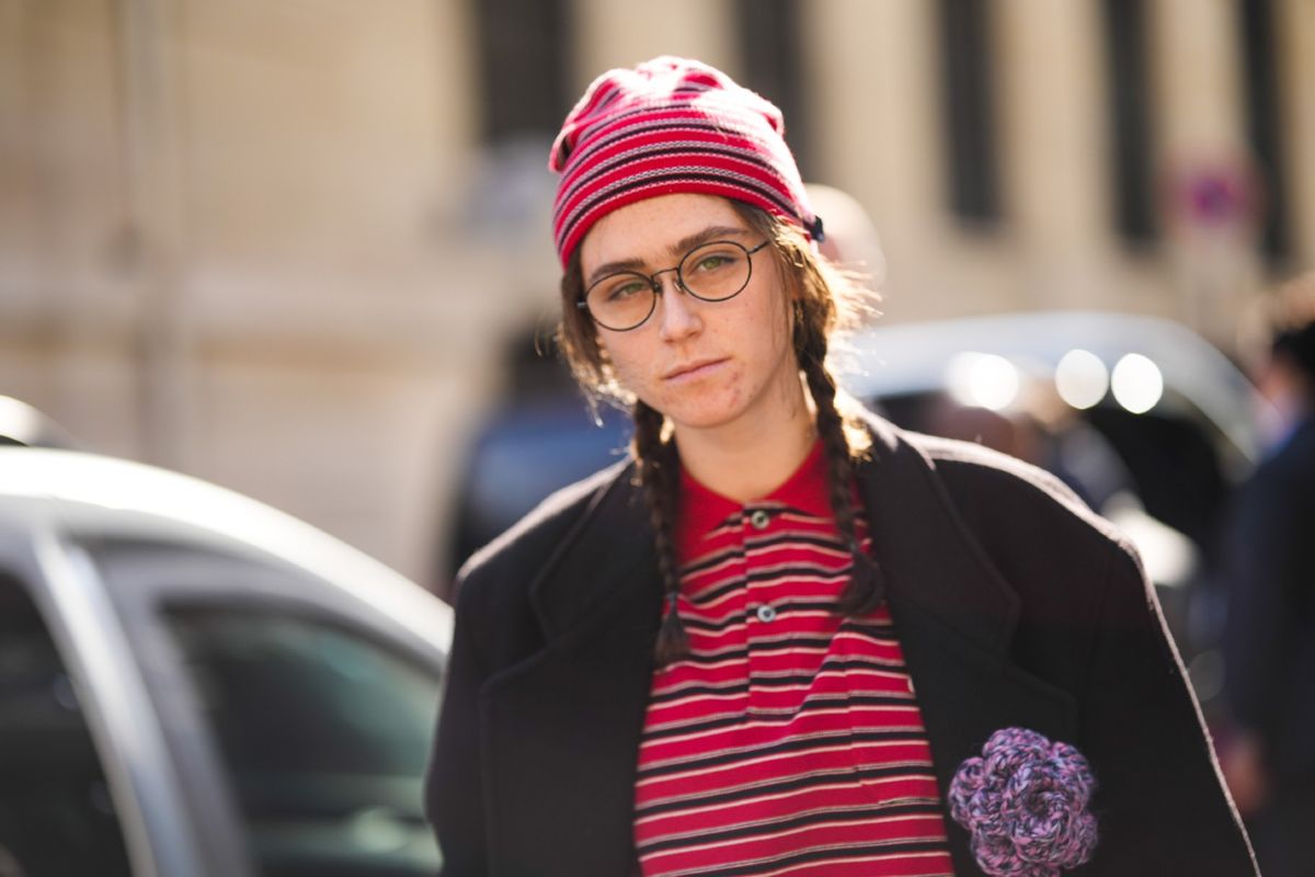 Ella Emhoff wears a red with black and white striped print pattern beanie, a matching red with black and white striped print pattern polo shirt, a black long coat with a purple flower brooch, black glasses, gray large pants, outside Dries Van Noten, during Paris Fashion Week - Womenswear Fall Winter 2023 2024, on March 01, 2023 in Paris, France.  (Edward Berthelot/Getty Images)