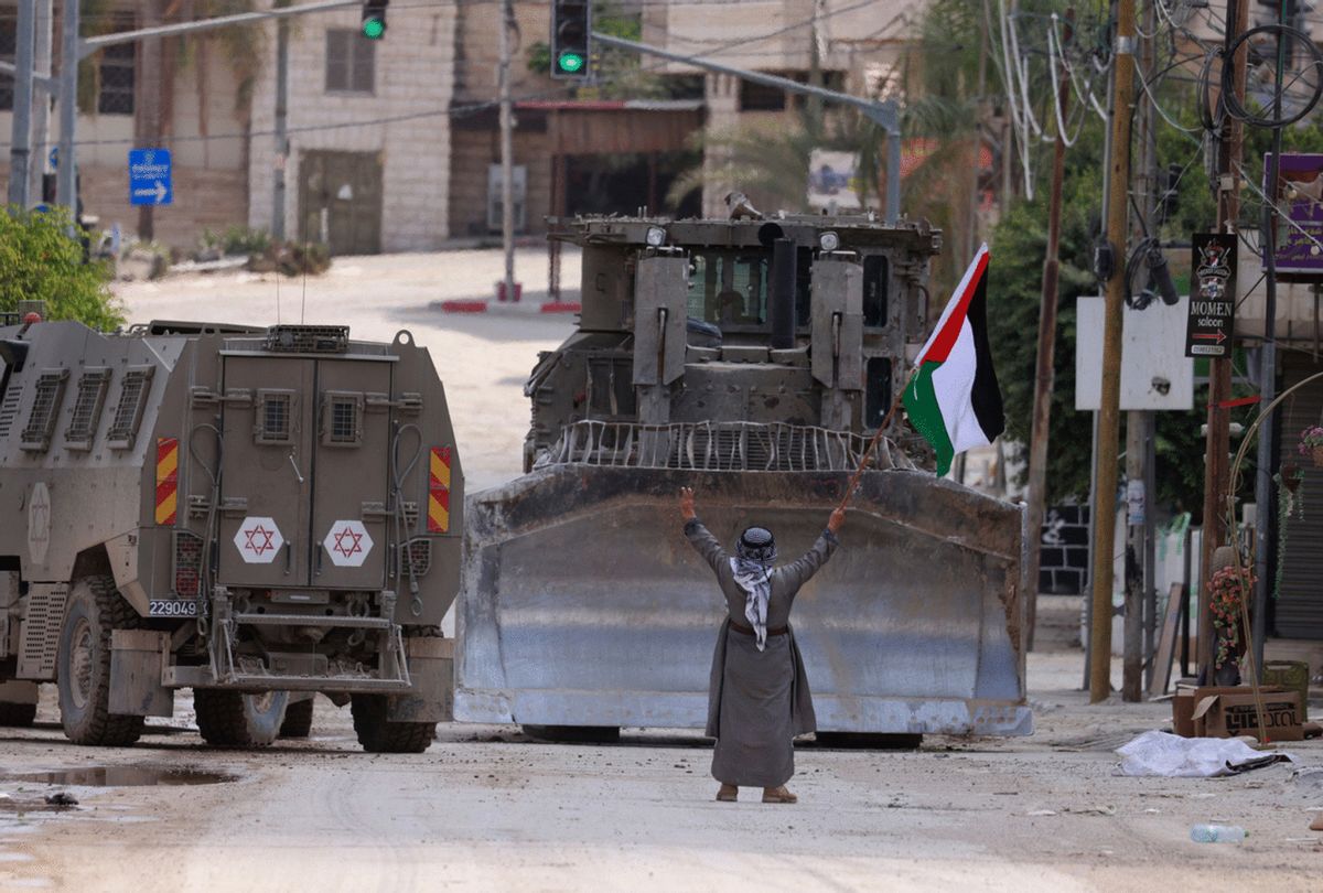 TOPSHOT - A Palestinian activist lifts a national flag and flashes the victory sign as Israeli armoured vehicles including a bulldozer drive on a street during a raid in Tulkarem on September 3, 2024, amid a large-scale military offensive launched a week earlier in the occupied West Bank. (Photo by Jaafar ASHTIYEH / AFP) (Photo by JAAFAR ASHTIYEH/AFP via Getty Images)