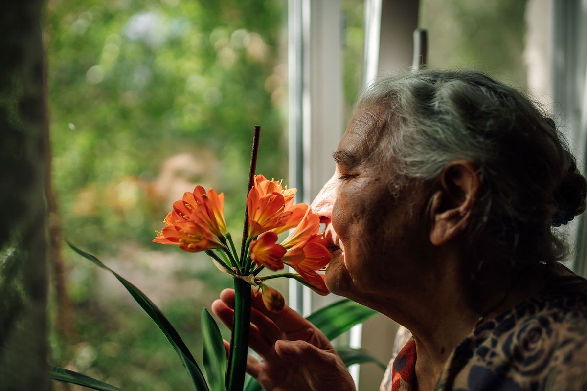 Elderly woman pauses by window to smell orange flowers. (Getty Images/Iuliia Burmistrova)