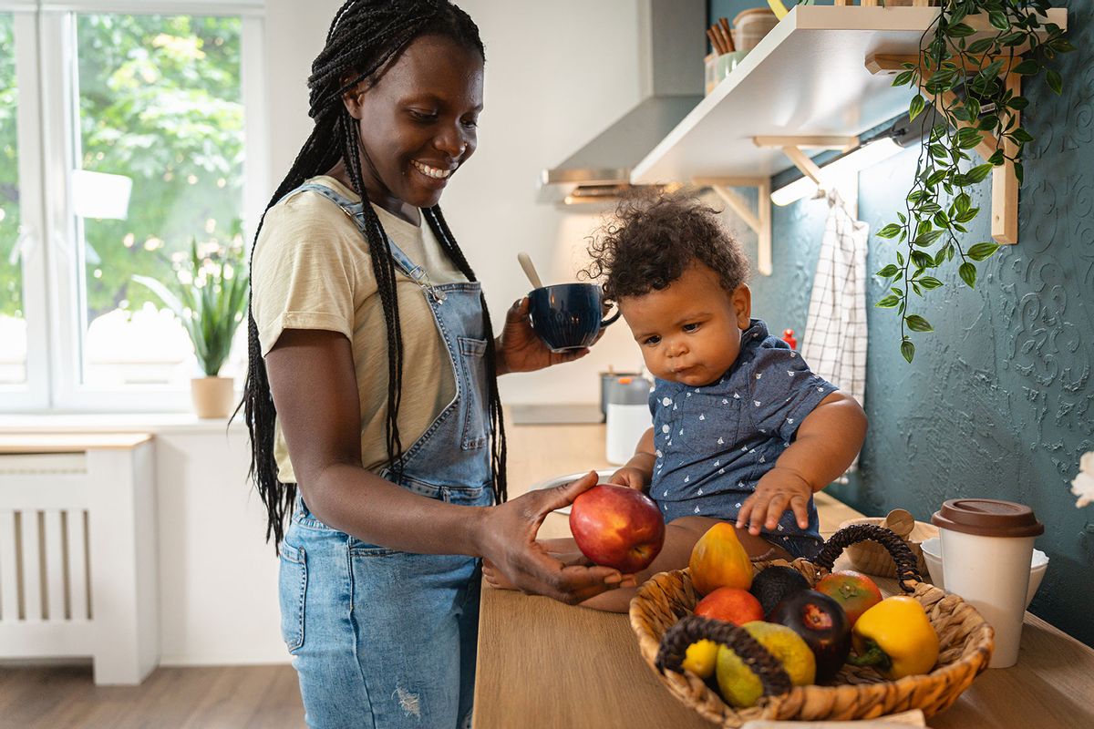 Playful baby boy sitting on the kitchen counter and playing with apple, while his mother has a coffee (Getty Images/FluxFactory)
