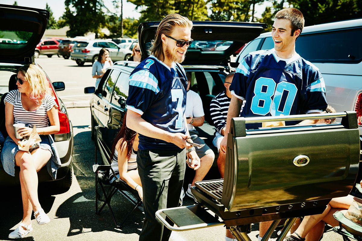 Smiling friends wearing team jerseys barbecuing at tailgating party in football stadium parking lot (Getty Images/Thomas Barwick)