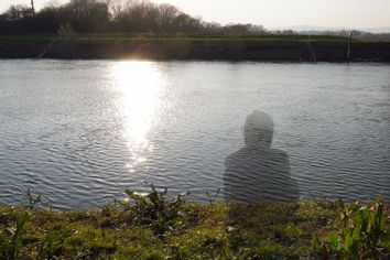 A transparent hooded figure sitting next to a river on a sunny afternoon