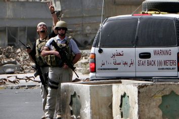 A private military contractor gestures to colleagues flying ovehead in a helicopter as they secure the scene of a roadside bomb attack in Baghdad, Iraq