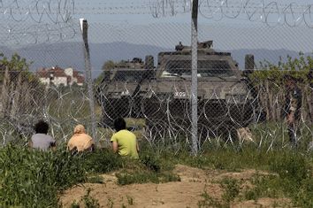 Migrants sit next to a border fence as Macedonian police stand guard at a makeshift camp for refugees and migrants at the Greek-Macedonian border near the village of Idomeni