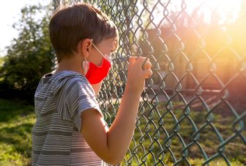 Serious upset little boy wearing a protective face mask looking through a school fence shaking it