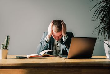 Worried businessman in dark suit sitting at office desk with laptop and notepad being overloaded with work