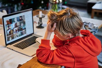 An elementary school student sits in front of a screen at home, using a laptop to participate in online classes with a teacher.