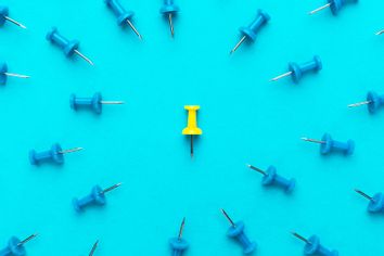 Yellow push pin in the center of blue push pins with their spike turned to the yellow one
