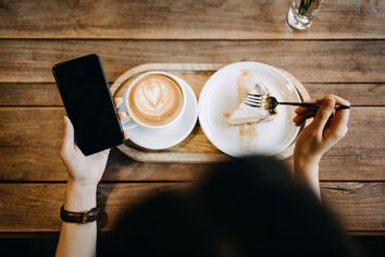 Woman holding her smartphone while enjoying a cup of coffee with cake in a coffee shop