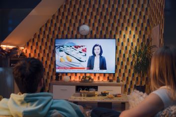 Couple sitting on sofa in living room with snacks and soda, watching TV news about medicine, new drugs and treatment