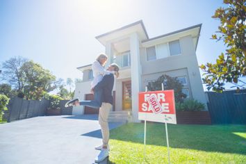 Couple celebrating in front of their new home with a real estate for sale sign saying sold