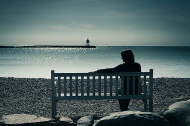 Woman in silhouette, sitting on a bench in a moody landscape at water's edge