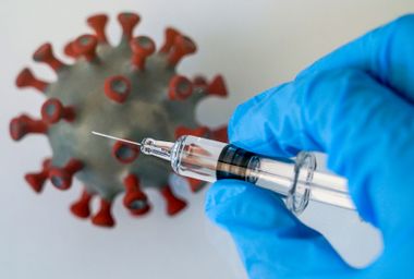 A vaccine syringe is held over a model of a coronavirus