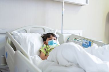 Little child patient with protective face mask lying on bed at hospital