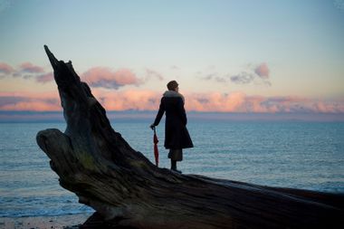 Woman leaning on umbrella standing on large driftwood tree trunk on beach