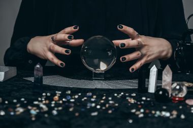 The hands of a young woman conjure over a transparent sphere surrounded by tarot cards, gems and other decor
