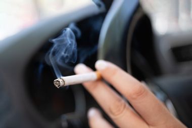 A woman sits in a car and holds a cigarette.