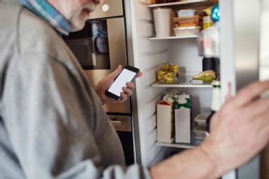 Senior man holding phone while searching in refrigerator at home