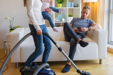 Woman with small child in her hands doing housekeeping while man sitting in couch and relaxing