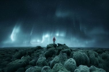 Distant silhouette of a person standing on rocks below Northern Lights